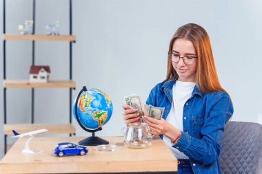 Genç kız masada otururken kumbarasından para sayar. Finansal okuryazarlık ve tasarruf kavramı. Banknotlarla şeffaf kumbara.