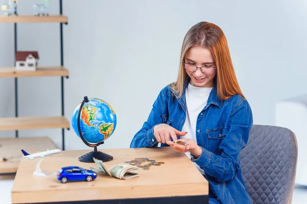 Hermosa chica caucásica cuenta sus ahorros para vacaciones en el extranjero. Mujer joven se sienta a la mesa con frasco de vidrio, dinero, globo, modelo de avión y coche . —  Fotos de Stock