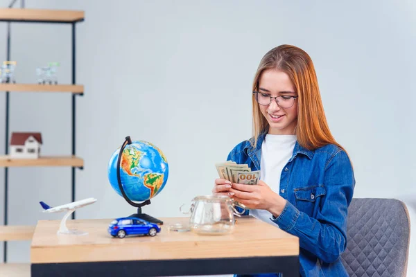 Девушка берет деньги из стеклянной банки на деревянном столе с глобусом, игрушечной машиной и самолетом на фоне белой комнаты. Крупный план . — стоковое фото