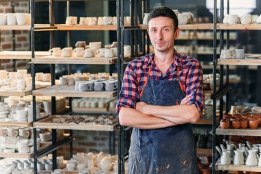 Önlük giyen ve arka planda seramik dolu raflar ile çömlek atölyesinde kamera yada bakan elleri geçiş ile çekici erkek çömlekçi.