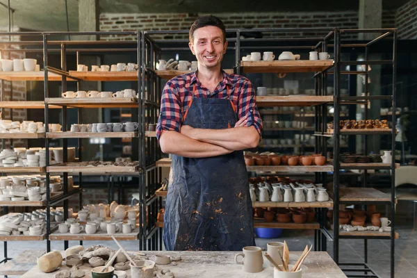 Önlük giyen ve arka planda seramik dolu raflar ile çömlek atölyesinde kamera yada bakan elleri geçiş ile çekici erkek çömlekçi.