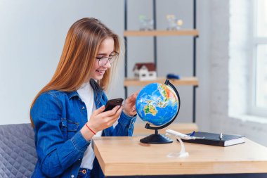 Genç beyaz kadın masaya oturur ve tatil için ülke seçmek ve not defterine notlar yapmak için dünya döndürür.