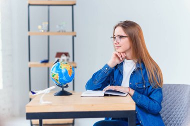 Genç kız beyaz oturma odasında okurken tatil hakkında rüyalar. Dünya ile coğrafya eğitimi. Denim giysiler içinde Sevimli kız öğrenci ev ödevi yapar.