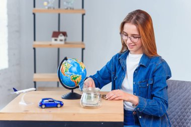 Genç kız bir masada otururken bir kumbara dan para sayar.