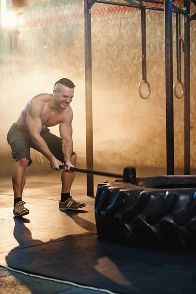 Strong young man hitting wheel tire using hammer in the gym.