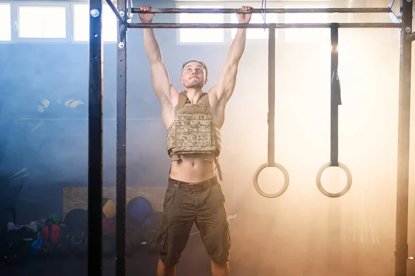 Jovem homem de esportes com colete pesado faz pull ups na barra transversal no ginásio cheio de fumaça . — Fotografia de Stock