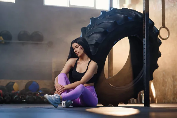 Mujer deportiva cansada descansando después del entrenamiento mientras está sentada en el suelo cerca del neumático de rueda en el gimnasio . — Foto de Stock