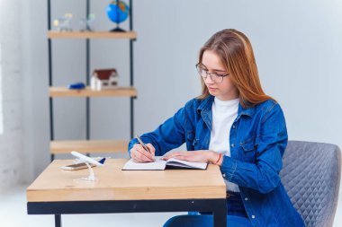 Ofiste denim kıyafetler giyen güzel genç kadın, not defterine bazı notlar yapmak.