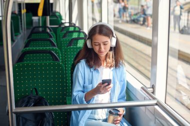 Güzel kadın modern şehir tramvay hareket ederken kulaklık, akıllı telefon ve kahve fincan ile oturur. toplu taşıma da gezi zevk.