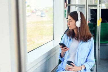 Toplu taşıma araçlarını kullanan genç kadın modern otobüste akıllı telefon ve beyaz kulaklıkla oturuyor.