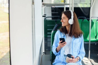 Genç kadın modern şehir otobüsünde oturur, müzik dinler, kahve içer ve pencereden dışarı bakar.