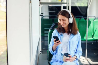Toplu taşıma araçlarını kullanan genç kadın modern otobüste akıllı telefon ve beyaz kulaklıkla oturuyor.