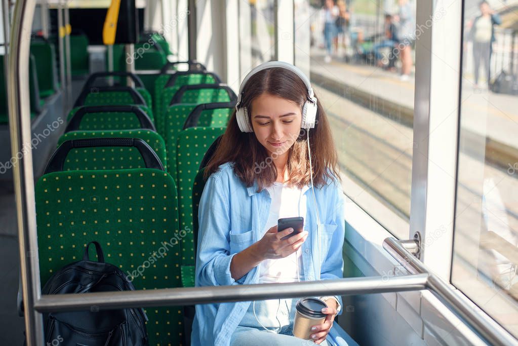 Pretty woman sits with headphones, smartphone and cup of coffee while moving in modern city tram. enjoying the trip at the public transport.