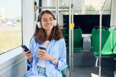 Genç kadın modern şehir otobüsünde oturur, müzik dinler, kahve içer ve pencereden dışarı bakar.