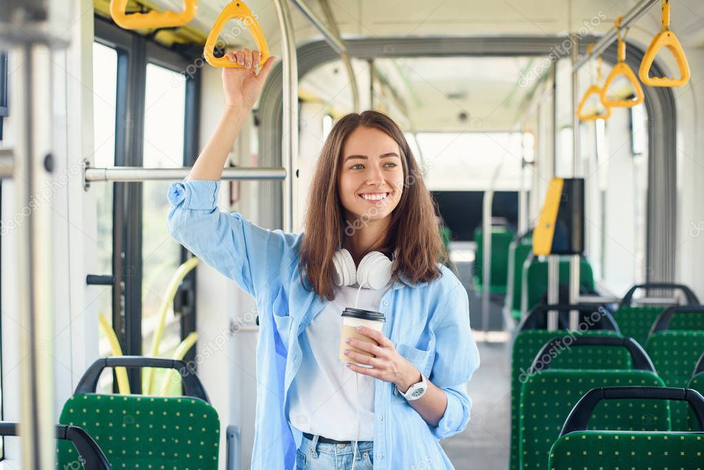 Stylish woman in blue shirt enjoying trip in the modern tram or bus, stands with cup of coffee in the public transport.