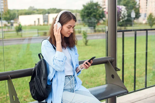 Teen girl listens to the music by white headphones in a public transport station while she waiting for tram.