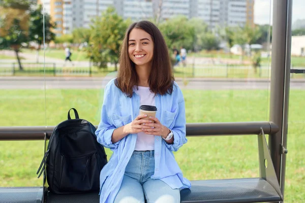 Kouzelná dívka alternativců čeká na autobus nebo tramvaj na nádraží veřejné dopravy ráno s šálkem kávy a používá chytrý telefon a Internet pro sledování přenosu přes App. — Stock fotografie