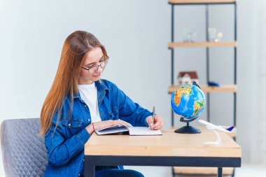 Öğrenci kız beyaz oturma odasında okurken not defterine yazıyor. Dünya ile coğrafya eğitimi. Denim giysiler içinde Sevimli kız öğrenci hometask yapar.