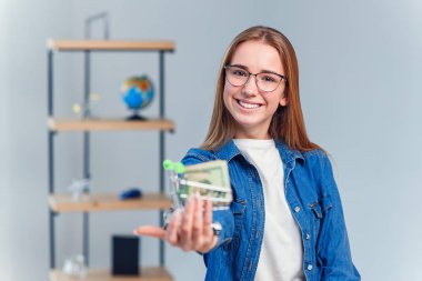 Denim gömlek ve gözlük genç güzel kadın Abd doları dolu küçük bir alışveriş sepeti tutar. Alışveriş, para tasarrufu kavramı.