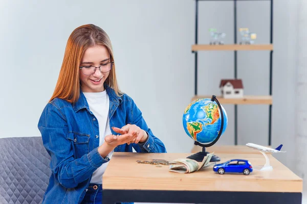 Güzel beyaz kız, birikimlerini yabancı tatillere sayar. Genç kadın cam kavanoz, para, küre, hava uçağı ve araba modeli ile masada oturur.