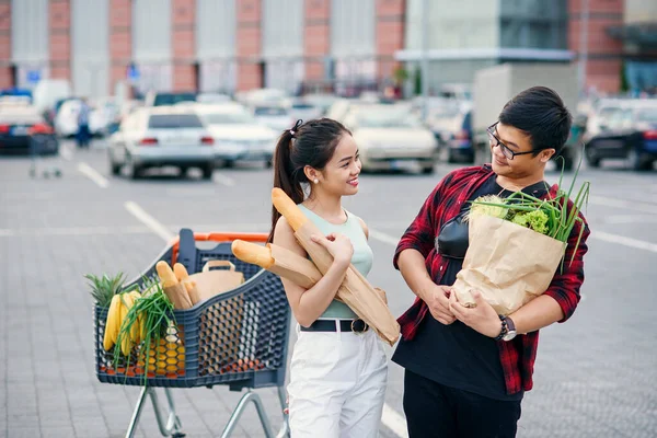 Piacevole donna asiatica e bello vietnamita giovane guardare l'un l'altro tenendo sacchetti di carta eco con cibo biologico sano nelle mani vicino al centro commerciale. Shopping in famiglia nel fine settimana . — Foto Stock