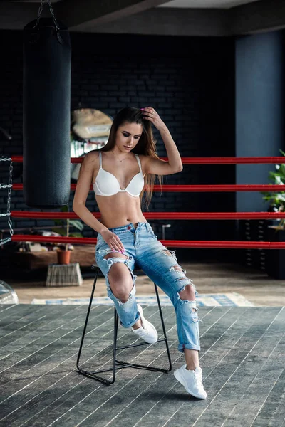 Prachtig meisje in lingerie en denim broek op boksring, zit op stoel in moderne loft gym. — Stockfoto