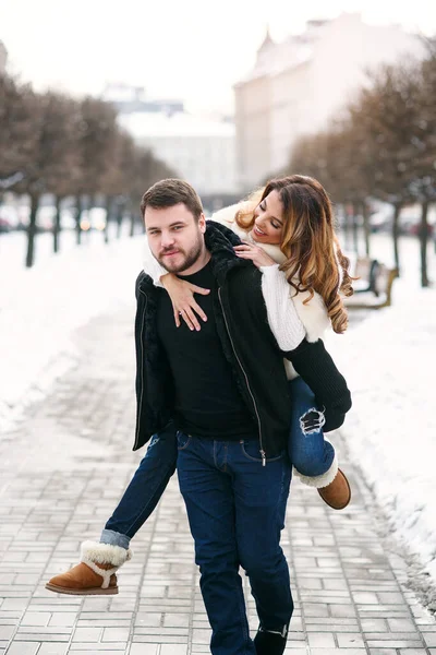 युवा स्वस्थ आदमी सर्दियों की बर्फीली सड़क की पृष्ठभूमि पर हाथों पर अपनी सुंदर प्रेमिका रखता है। नए साल और क्रिसमस अवधारणा . — स्टॉक फ़ोटो, इमेज