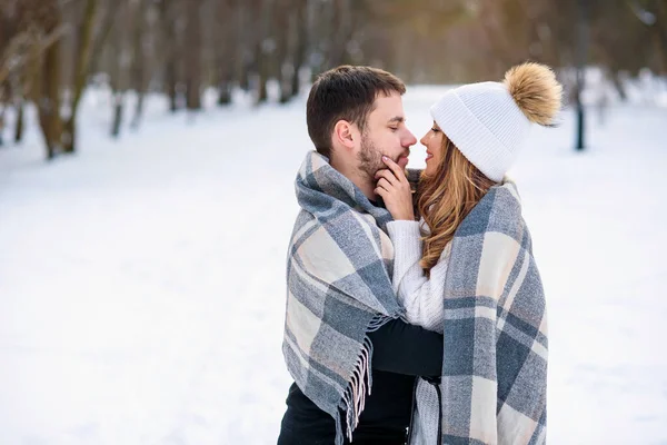 Belle fille en chapeau blanc chaud et son petit ami s'embrassant et couvert de carreaux à carreaux tout en marchant dans un parc d'hiver enneigé . — Photo