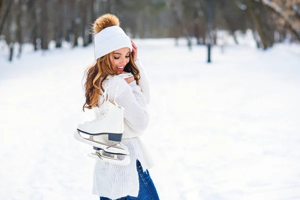 在冬季的雪地公园里，漂亮的女人穿着白色的毛衣，戴着一顶帽子，背上穿着冰鞋. — 图库照片