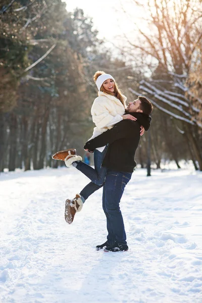 Молодая стильная влюбленная пара веселится во время прогулки по снежному зимнему парку. Красивый молодой человек держит свою красивую девушку на руках . — стоковое фото