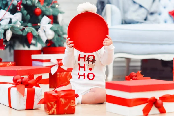 Affascinante bambino seduto tra i regali di festa in una stanza accogliente e coperto il viso con una copertina di carta circolare rossa dalla scatola . — Foto Stock