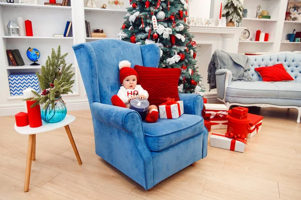 Menino pequeno bonito na roupa do Natal senta-se na cadeira grande azul e segurando a caixa de presente em suas mãos . — Fotografia de Stock