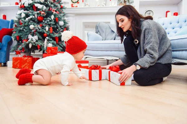 Adorável sorrindo bebê rastejando no parquete para os presentes de Natal perto de sua mãe, que se senta no chão . — Fotografia de Stock