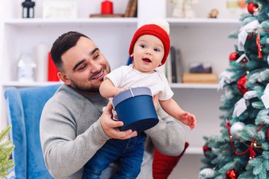 Genç baba, elinde komik bir bebekle rahat koltukta oturuyor ve Noel 'i kutlamak için muhteşem dekore edilmiş odasında ona hediye kutusu veriyor..
