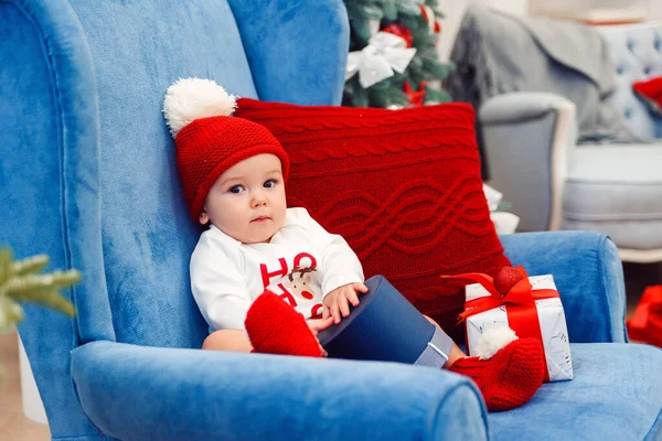 Beau petit garçon en vêtements de Noël est assis dans une grande chaise bleue et tenant boîte cadeau dans ses mains . — Photo