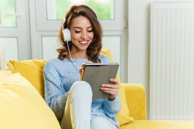 Genç ve güzel bir kız modern sarı koltukta oturup müzik dinliyor ve evde tablet kullanıyor..