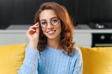 25 yaşında, yumuşak mavi süveterli, gözlüklü güzel bir kız kameraya bakıyor ve şık mutfağın arka planında harika bir gülümseme var..