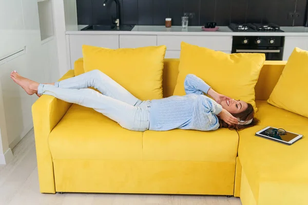 Menina adolescente loira bonita em fones de ouvido está ouvindo música, olhando para a câmera e sorrindo enquanto deitada de costas, pernas para cima, no sofá em casa — Fotografia de Stock