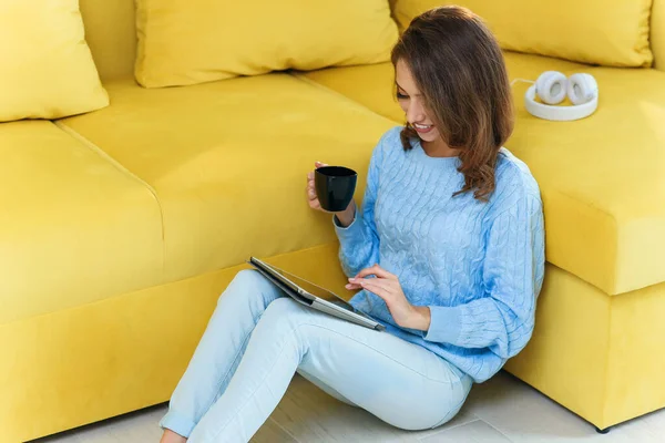 Encantadora mulher exuberante segurando seu ipad nas mãos, sentado no chão perto do sofá verde claro e desfrutando de café na xícara na cozinha contemporânea — Fotografia de Stock