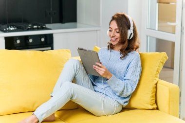 Kahverengi saçlı ve şık elbiseli muhteşem neşeli kadın mutfak kanepesinde dinleniyor ve kulaklıkla müzik dinlemek için dizüstü bilgisayar kullanıyor..