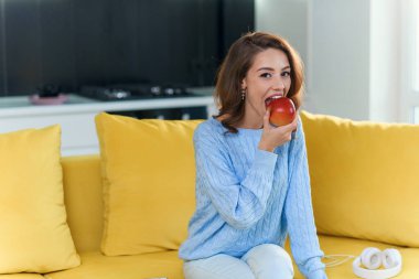 Güzel genç bir kadın modern mutfakta sulu kırmızı elma yer ve kameraya hoş bir gülümsemeyle poz verir..