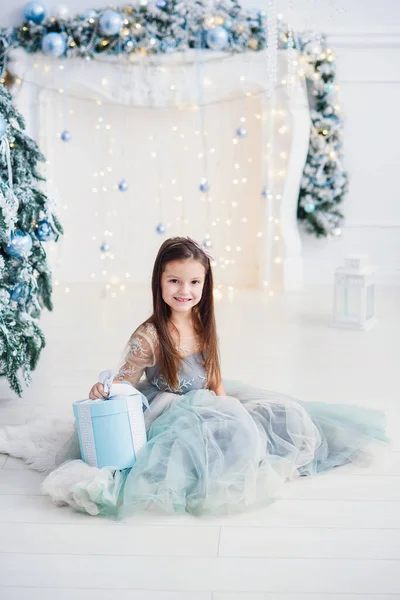 Carino bambina con scatola regalo in bel vestito si siede vicino all'albero di Natale sullo sfondo del camino . — Foto Stock