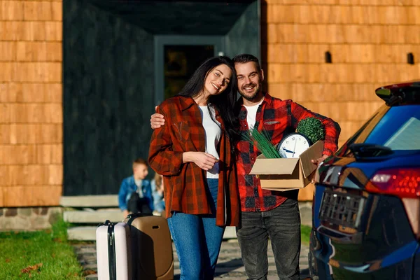 Adorável casal apaixonado sorrindo e sentindo felicidade ao ficar perto da casa moderna com caixa de papelão com relógio e plantas verdes durante a mudança para a nova casa . — Fotografia de Stock