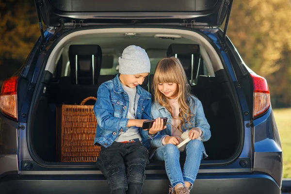 Movimento lento de loira menina de 12 anos e bonito menino de 10 anos em jeans roupas que se sentam no porta-malas e olhando em seu smartphone — Fotografia de Stock
