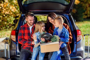 Sevimli, genç ebeveynler ve şirin çocukları bagajda oturuyorlar ve yeni bir daireye taşınırken ellerinde bitki ve diğer ev eşyalarıyla birlikte kutu tutuyorlar..