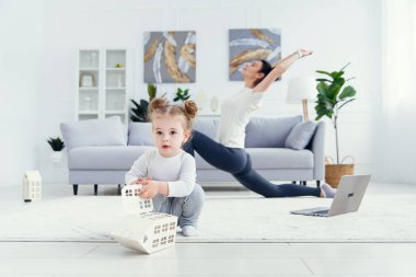 Sportif, sağlıklı annesi arka planda spor yaparken ve yoga egzersizleri yaparken evde oynayan komik bir bebek..