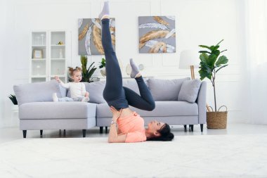 Mutlu anne, küçük kızı evde oynarken yoga pozisyonunda sabah egzersizi yapıyor. Genç sevimli anne meditasyon yaparken eğleniyor. Stresten uzak bir hafta sonu bebeğiyle rahatlıyor..