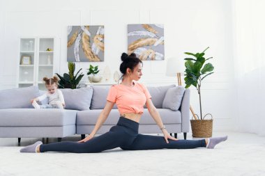 Mutlu anne, küçük kızı evde oynarken yoga pozisyonunda sabah egzersizi yapıyor. Genç sevimli anne meditasyon yaparken eğleniyor. Stresten uzak bir hafta sonu bebeğiyle rahatlıyor..
