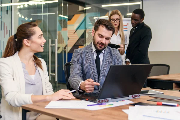Gemeinsame Arbeit zweier selbstbewusster Geschäftsleute im formellen Anzug, die mit Laptop im Büro arbeiten, mit ihren Kollegen im Hintergrund. — Stockfoto