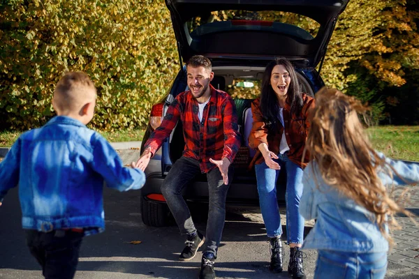Jovens pais sentados no porta-malas dos carros e pegam para abraçar seu filho e filha felizes que correm para eles . — Fotografia de Stock